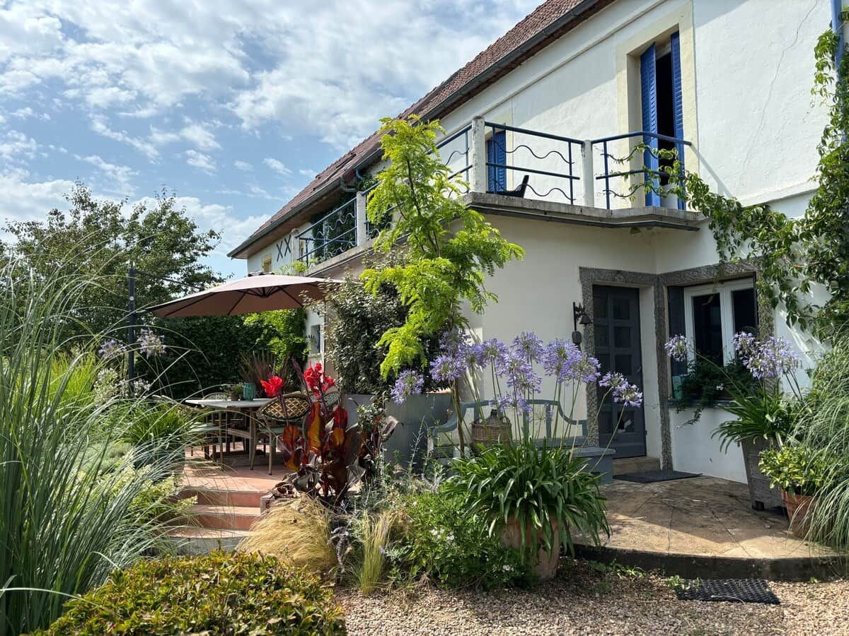 Une maison à deux étages avec un balcon, une terrasse dotée d’une table et de chaises, ainsi que diverses plantes et fleurs.