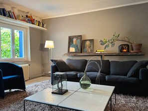 A living room with a sofa, a coffee table, and a lamp. There are bookshelves and a window with a view.