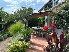 Une terrasse avec une table et des chaises, entourée de plantes en pot et agrémentée d’un grand parasol.