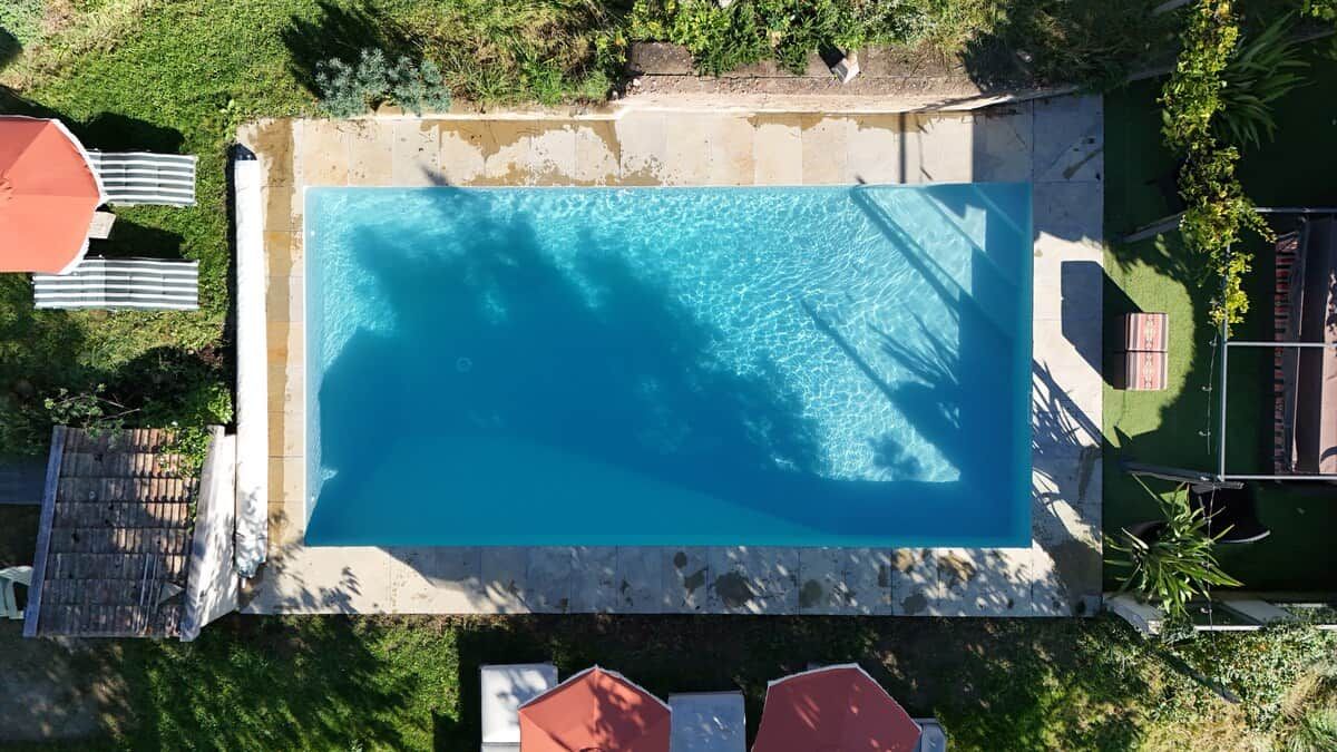 Une vue aérienne d’une piscine rectangulaire bordée de pierres, entourée d’une pelouse et de quelques arbres.