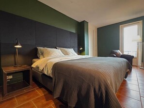 Un dormitorio con cama, mesita de noche, lámpara y vistas al exterior.