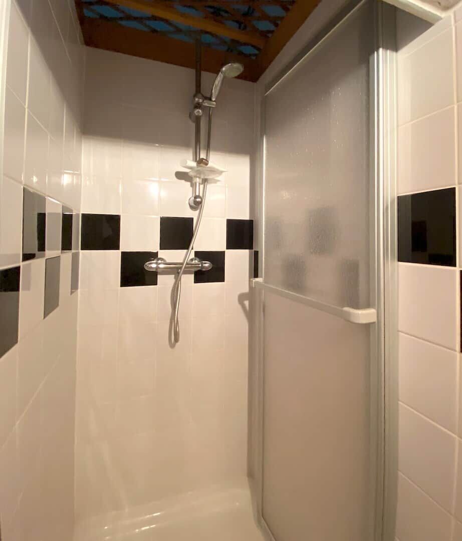Eine Dusche mit Glastür, weißen Fliesen mit schwarzen Akzenten und einer Holzbalkendecke.