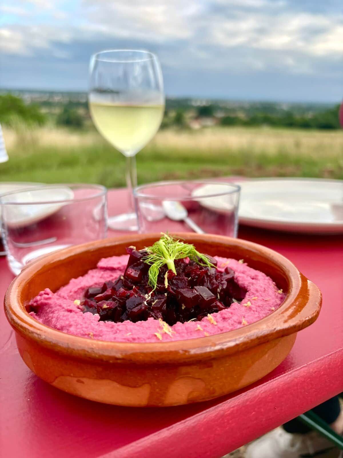 Una mesa con un cuenco de comida, una copa de vino blanco y una vista panorámica al fondo.