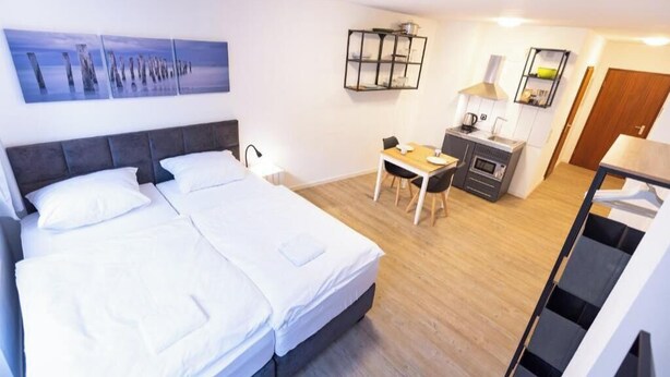 Habitación de hotel con cama, escritorio, silla, kitchenette pequeña y estante fijado a la pared.