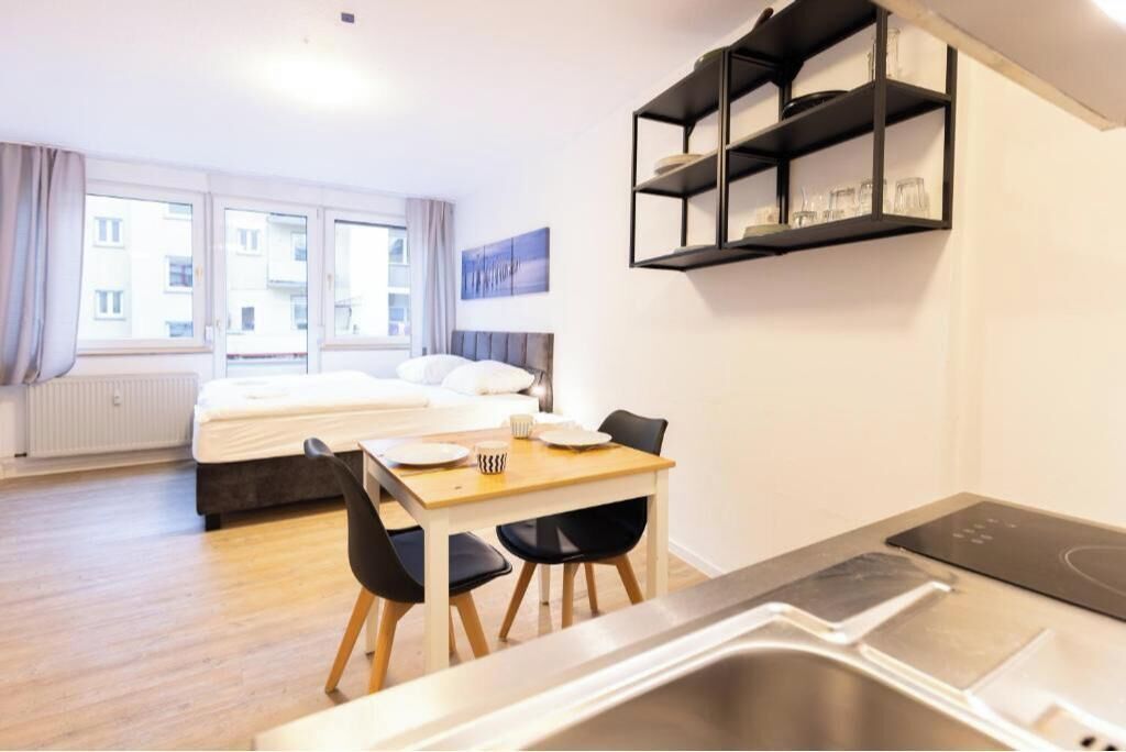 A compact hotel room with a bed, a dining area with a table and chairs, a built-in shelf with glasses, and a window with curtains.