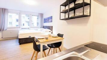 A compact hotel room with a bed, a dining area with a table and chairs, a built-in shelf with glasses, and a window with curtains.