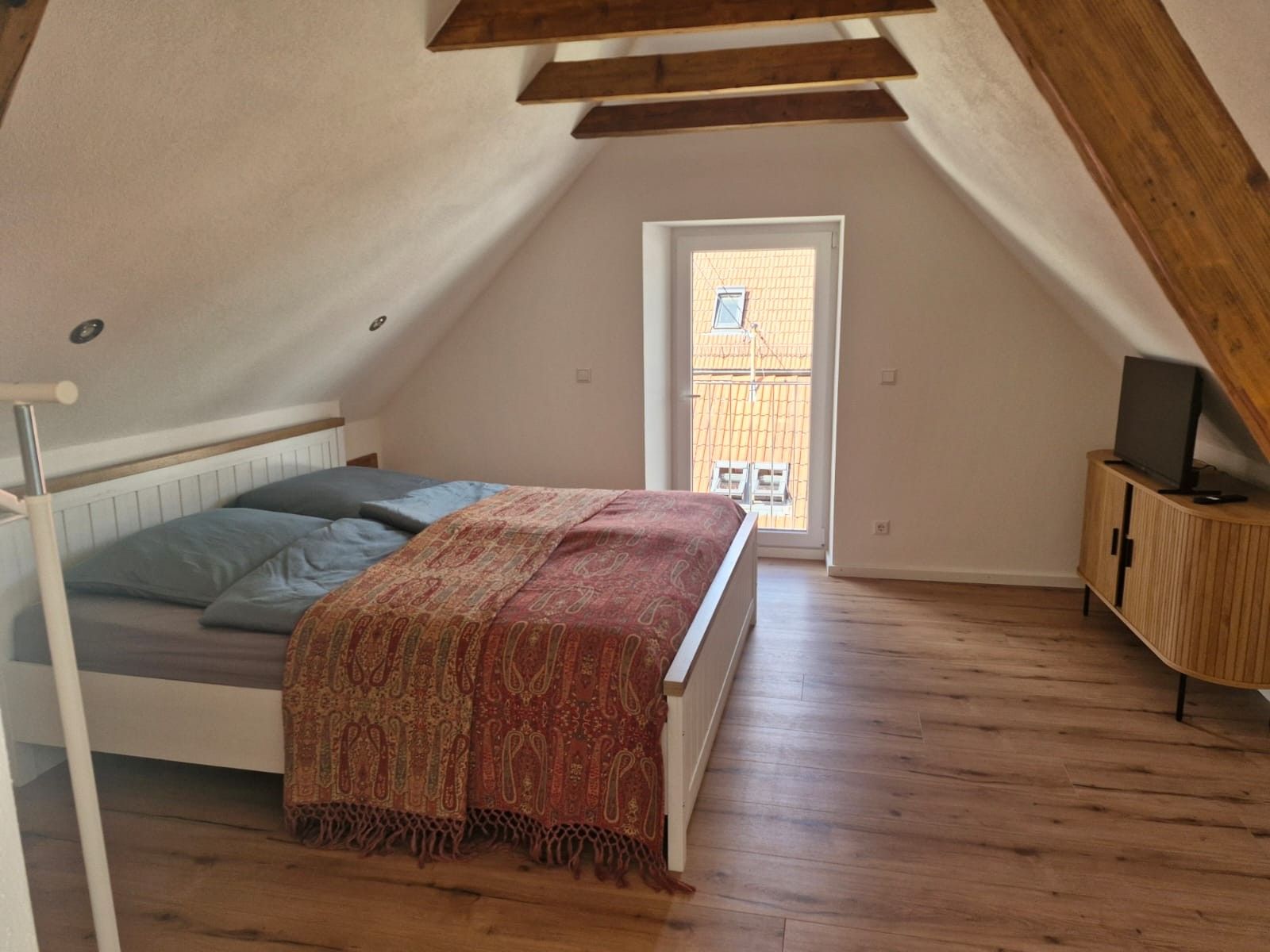 Ein Schlafzimmer mit schrägen Wänden, einem Bett mit gemustertem Überwurf, einem Holzschrank und einem Fenster mit Blick auf Gebäude.
