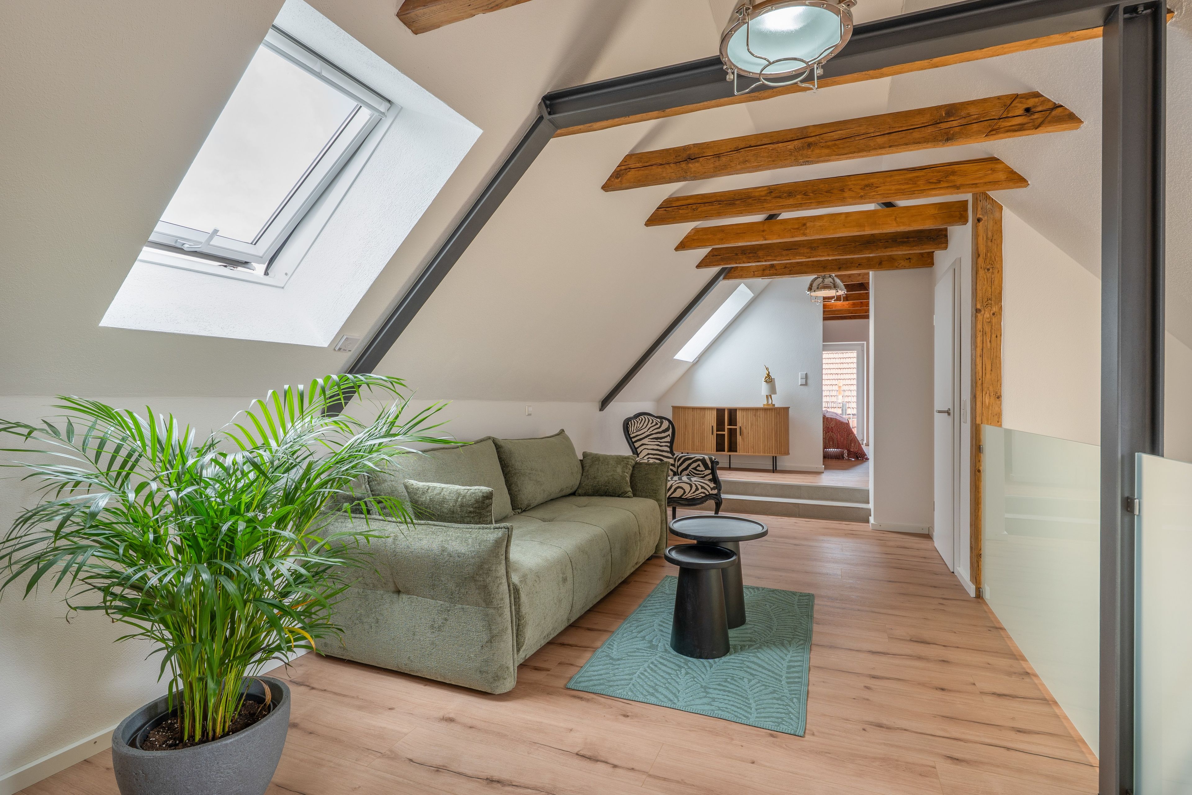 Ein Loft-Zimmer mit schrägem Dach, Holzbalken, einem grünen Sofa, einem kleinen runden Tisch und einem Blumentopf.