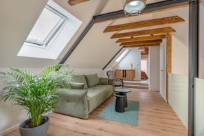 Une pièce de style « loft » avec un plafond en pente, des poutres en bois, un canapé vert, une petite table ronde et une plante en pot.