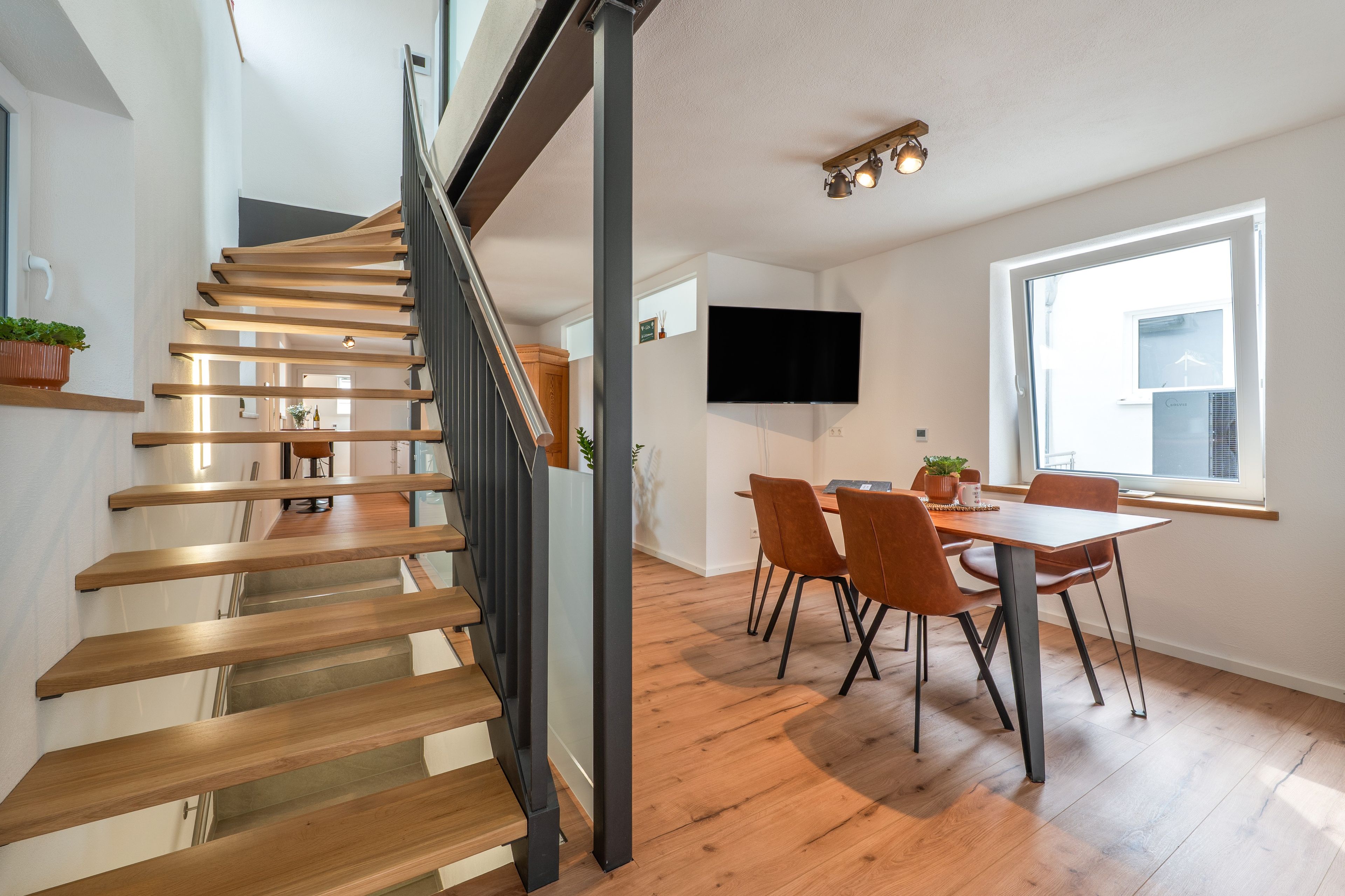 Een modern interieur met een houten trap, een eetgedeelte met stoelen, een flatscreen aan de muur en een groot raam dat voor daglicht zorgt.