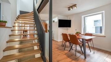 Een modern interieur met een houten trap, een eetgedeelte met stoelen, een flatscreen aan de muur en een groot raam dat voor daglicht zorgt.