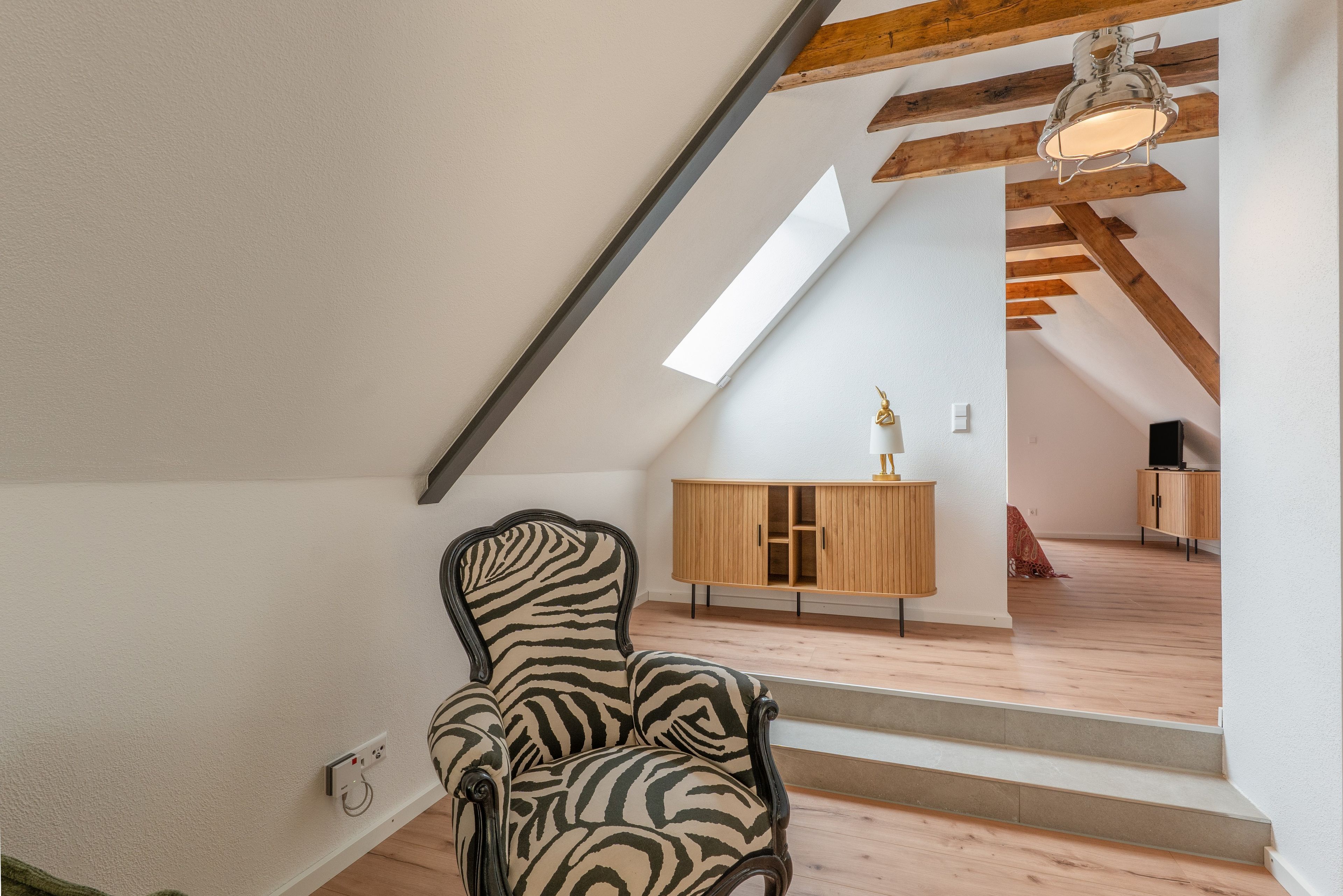 Een kamer met een stoel met een zebraprint, houten vloeren, een schuine plafond met zichtbare balken, een houten kast en een trap naar een hoger niveau.