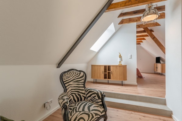 Une pièce avec un fauteuil à motifs zébrés, un plancher en bois, un plafond en pente avec des poutres apparentes, un meuble en bois et un escalier menant à un niveau supérieur.