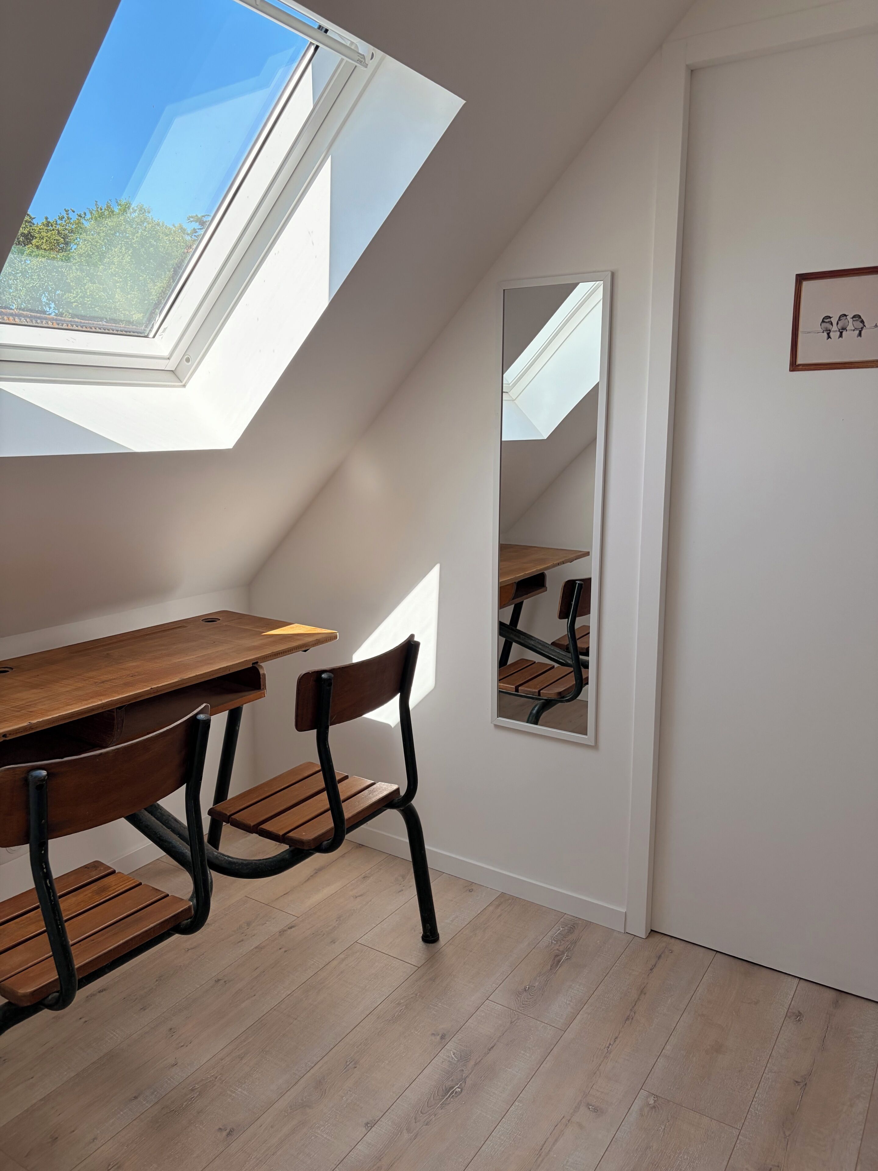 A room with a sloped ceiling, a wooden desk, two chairs, a mirror, and a framed picture on the wall.