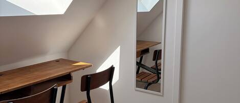 A room with a sloped ceiling, a wooden desk, two chairs, a mirror, and a framed picture on the wall.