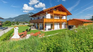 A two-story wooden chalet with a balcony, surrounded by lush greenery and mountains in the background. - Vrbo Property (Unterwössen)