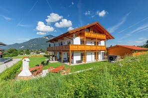 En tvåvånings chalet i trä med en balkong, omgiven av frodig grönska och berg i bakgrunden.