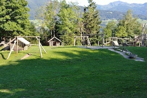 A playground with swings and a small wooden structure, surrounded by grass and trees, with mountains in the background. - Holiday apartment in Vomp with swimming pool (Vomp)