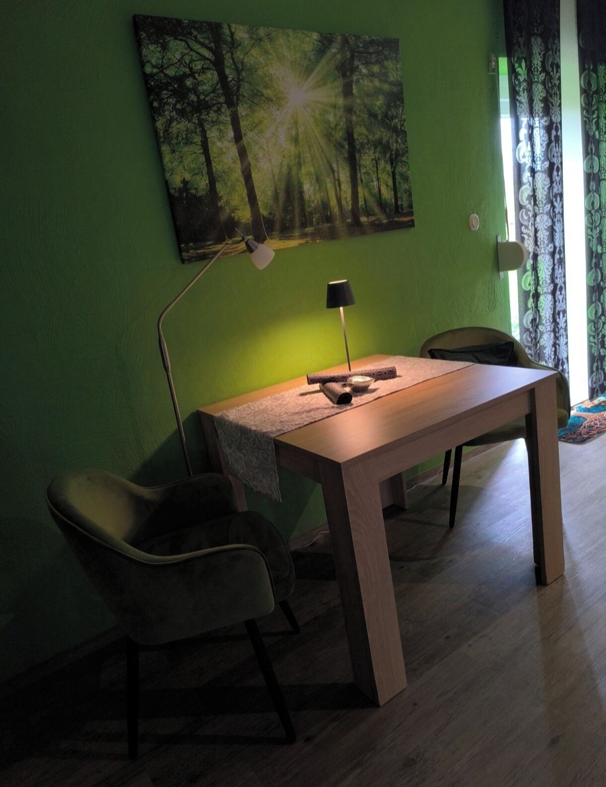 Ein Zimmer mit Schreibtisch aus Holz, zwei Stühlen, einer Lampe und einem Waldbild an der Wand.
