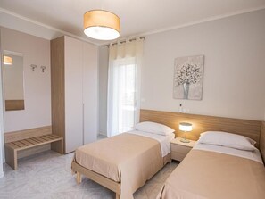 Una camera d'albergo con due letti, una panca, un comodino con una lampada, un quadro appeso al muro e uno specchio.