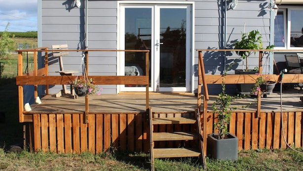 Um deck de madeira com banco e plantas em vasos, porta de correr de vidro e uma pequena mesa para área externa.