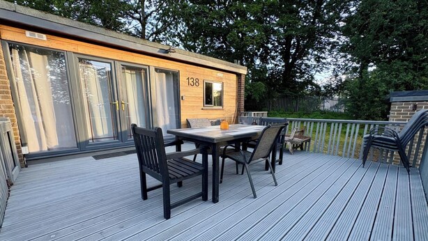 A deck with a table and chairs, a wooden building with the number 138, and a fenced area with trees. - Pet Friendly 2-bed Lodge, Sleeps 5, Pool & Parking (Caernarfon)