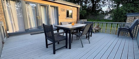 A deck with a table and chairs, a wooden building with the number 138, and a fenced area with trees.