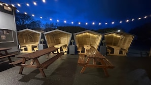 A series of wooden structures with benches and tables, illuminated by string lights, suggesting a cozy outdoor seating area. - Pet Friendly 2-bed Lodge, Sleeps 5, Pool & Parking (Caernarfon)