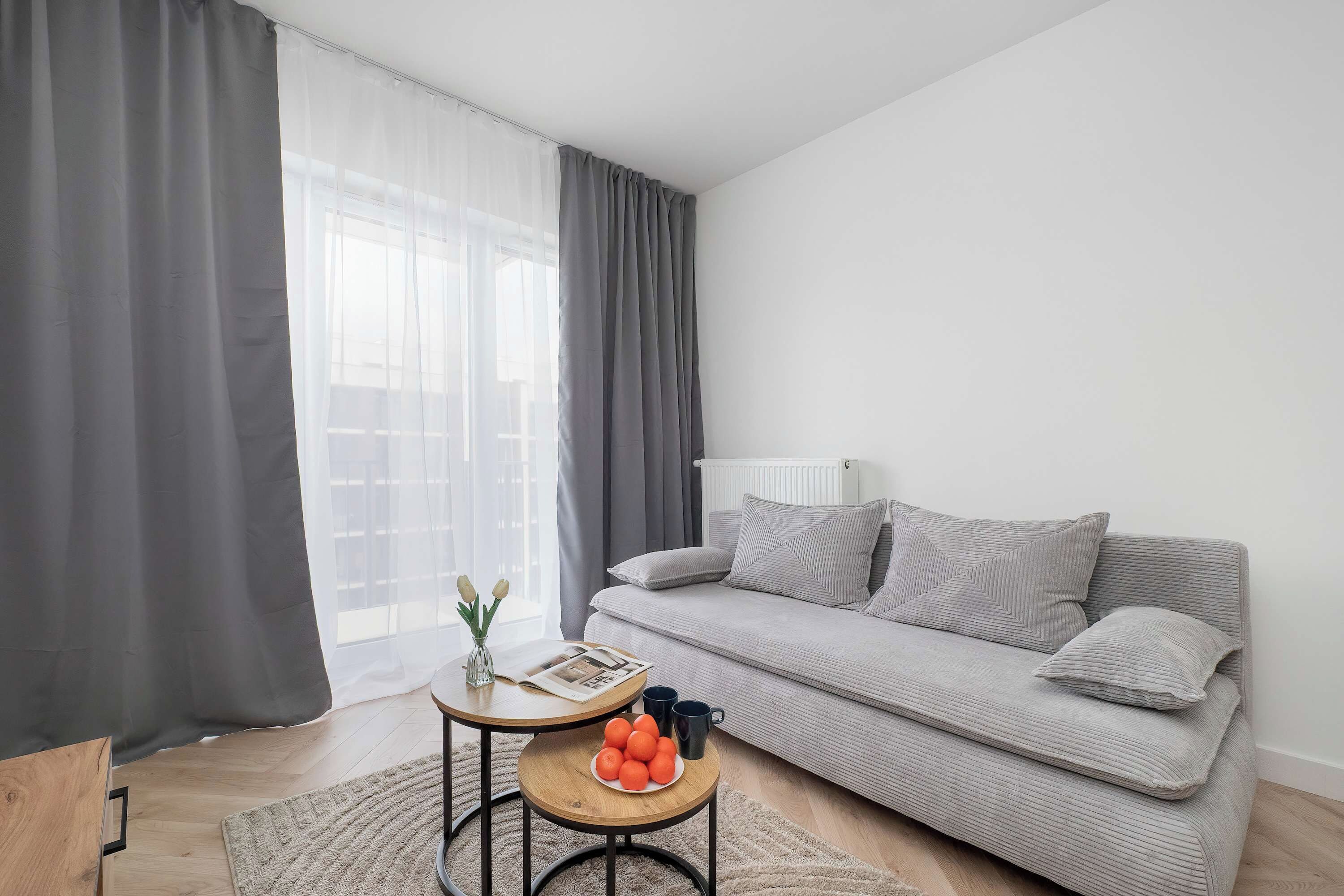 A modern living room with a grey sofa, a round wooden table with a vase and oranges, and a large window with sheer curtains.