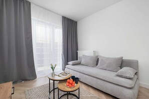 A modern living room with a grey sofa, a round wooden table with a vase and oranges, and a large window with sheer curtains. - Modern Apartment in the City Center | Balcony (Łódź)