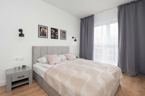 A modern bedroom with a bed, bedside tables, a nightstand, a wall-mounted lamp, and a window with curtains. - Modern Apartment in the City Center | Balcony (Łódź)
