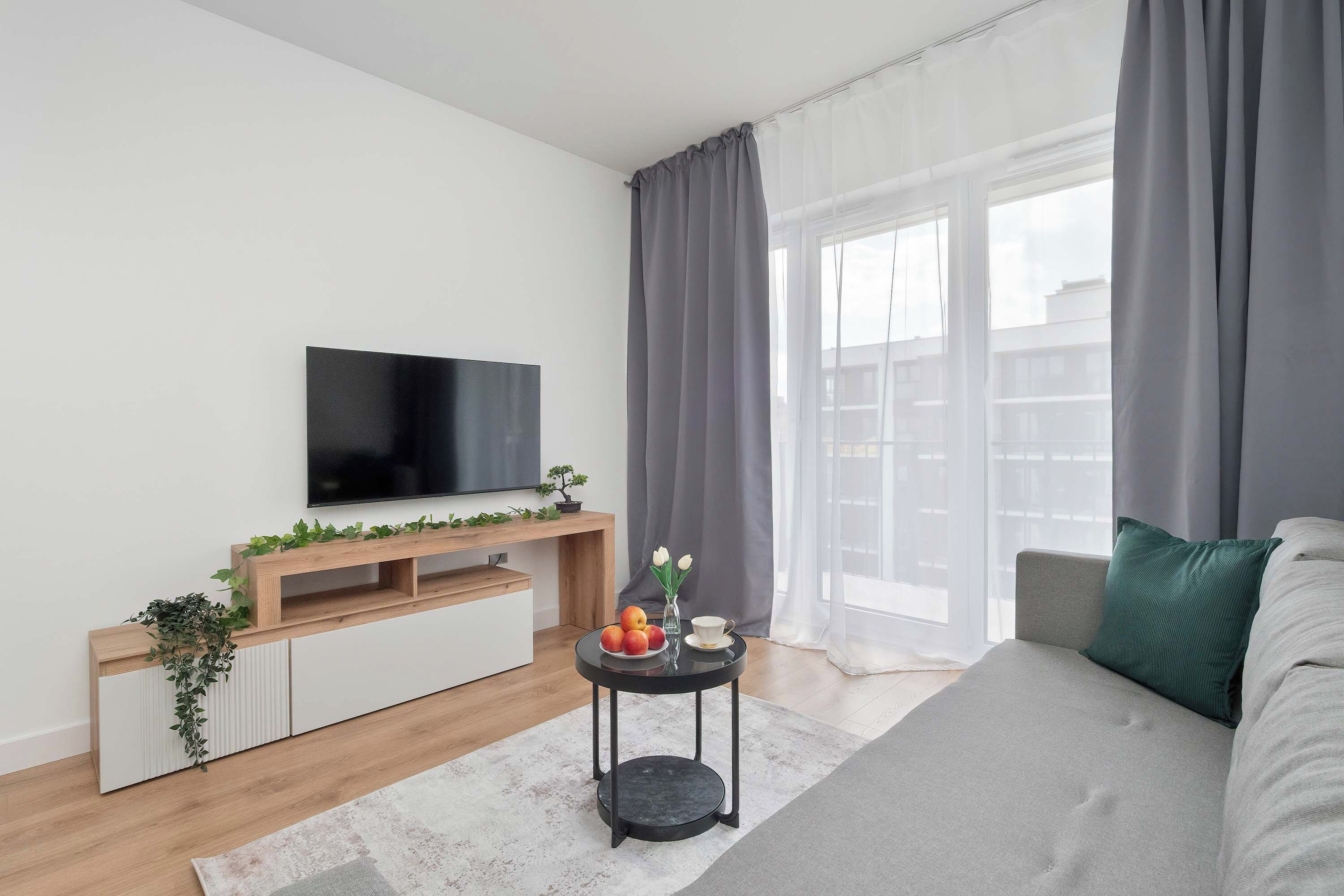Un salon moderne avec un téléviseur à écran plat, un meuble de rangement en bois, une petite table d’appoint ronde noire agrémentée d’un vase et de fruits, et un canapé gris avec un coussin vert.