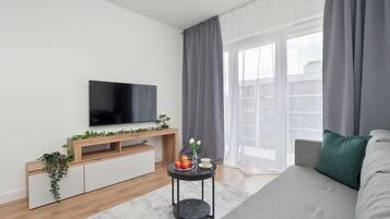 Uma sala de estar moderna com uma televisão de tela plana, um móvel de entretenimento de madeira, uma mesa lateral redonda preta com um vaso e frutas, e um sofá cinza com uma almofada verde.