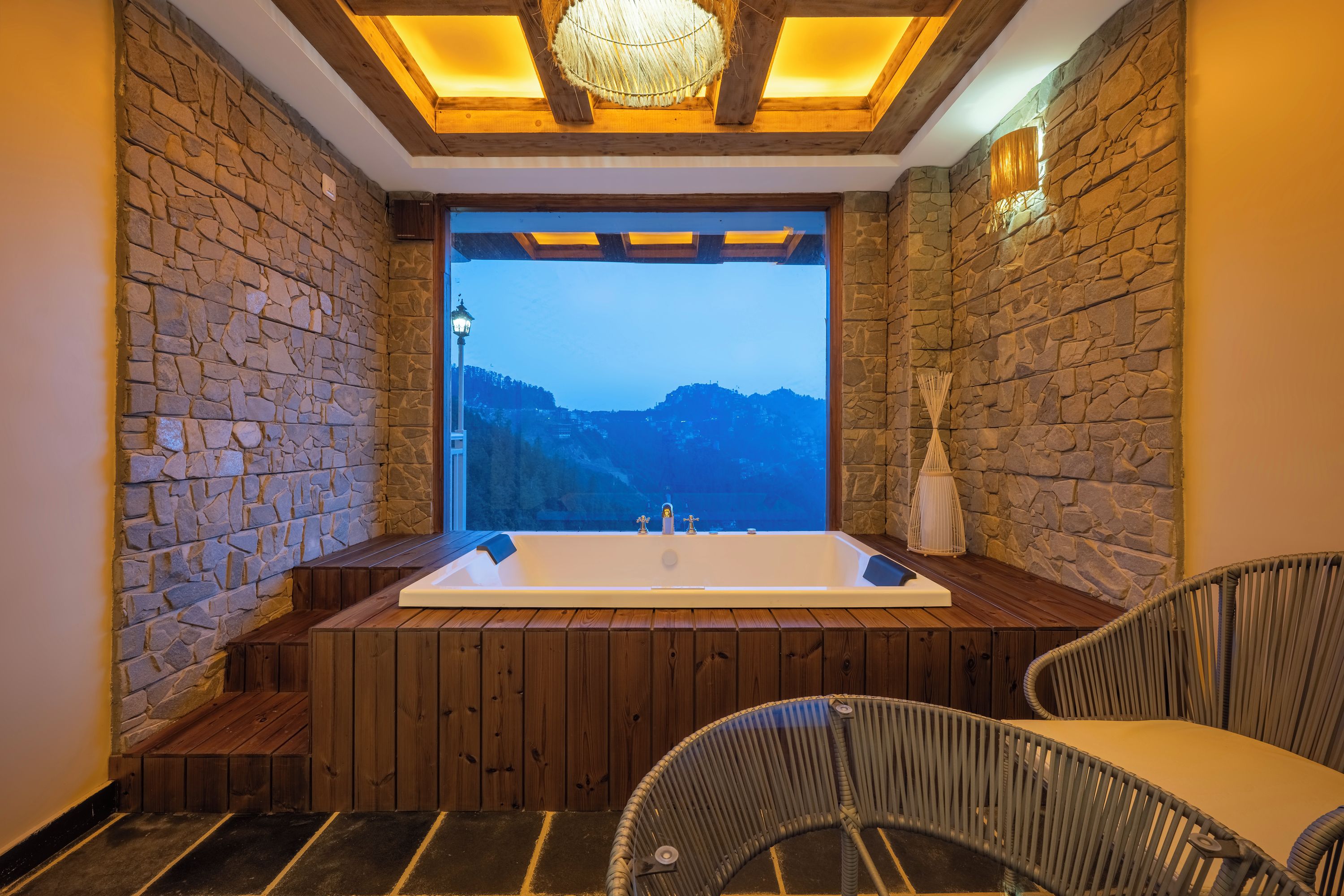Une salle de spa dotée d’une grande baignoire, de bancs en bois, de fauteuils en osier, de murs en pierre et offrant une vue sur une chaîne de montagnes.