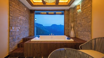 Une salle de spa dotée d’une grande baignoire, de bancs en bois, de fauteuils en osier, de murs en pierre et offrant une vue sur une chaîne de montagnes.