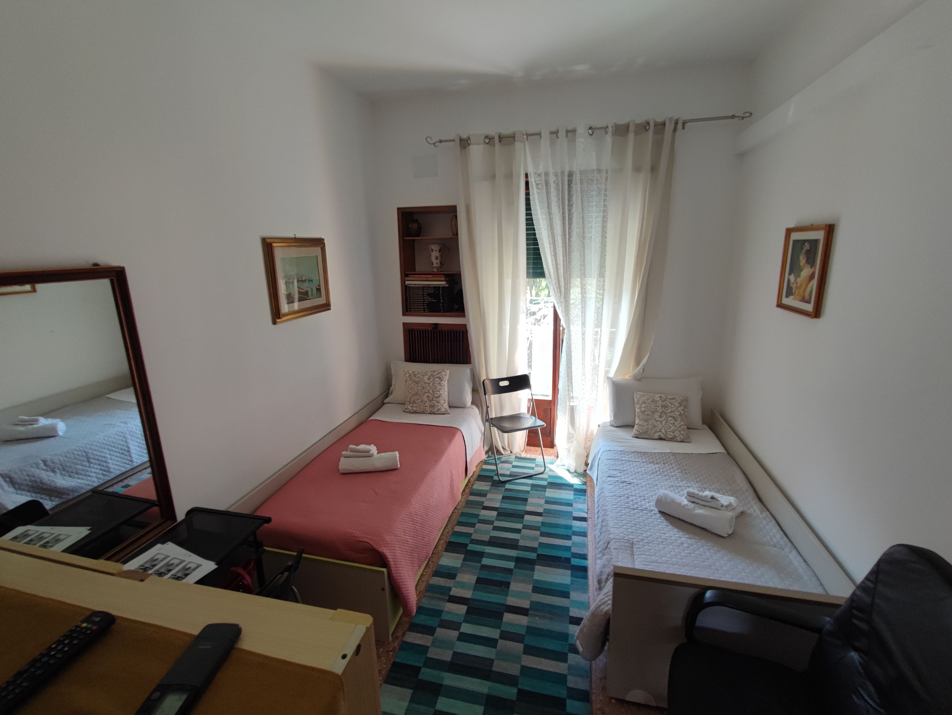 A hotel room with two beds, a desk with a computer, a chair, a bookshelf, and a window with curtains.