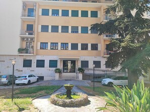 A multi-story residential building with a central entrance, a small garden with a fountain, and a parking area with cars.