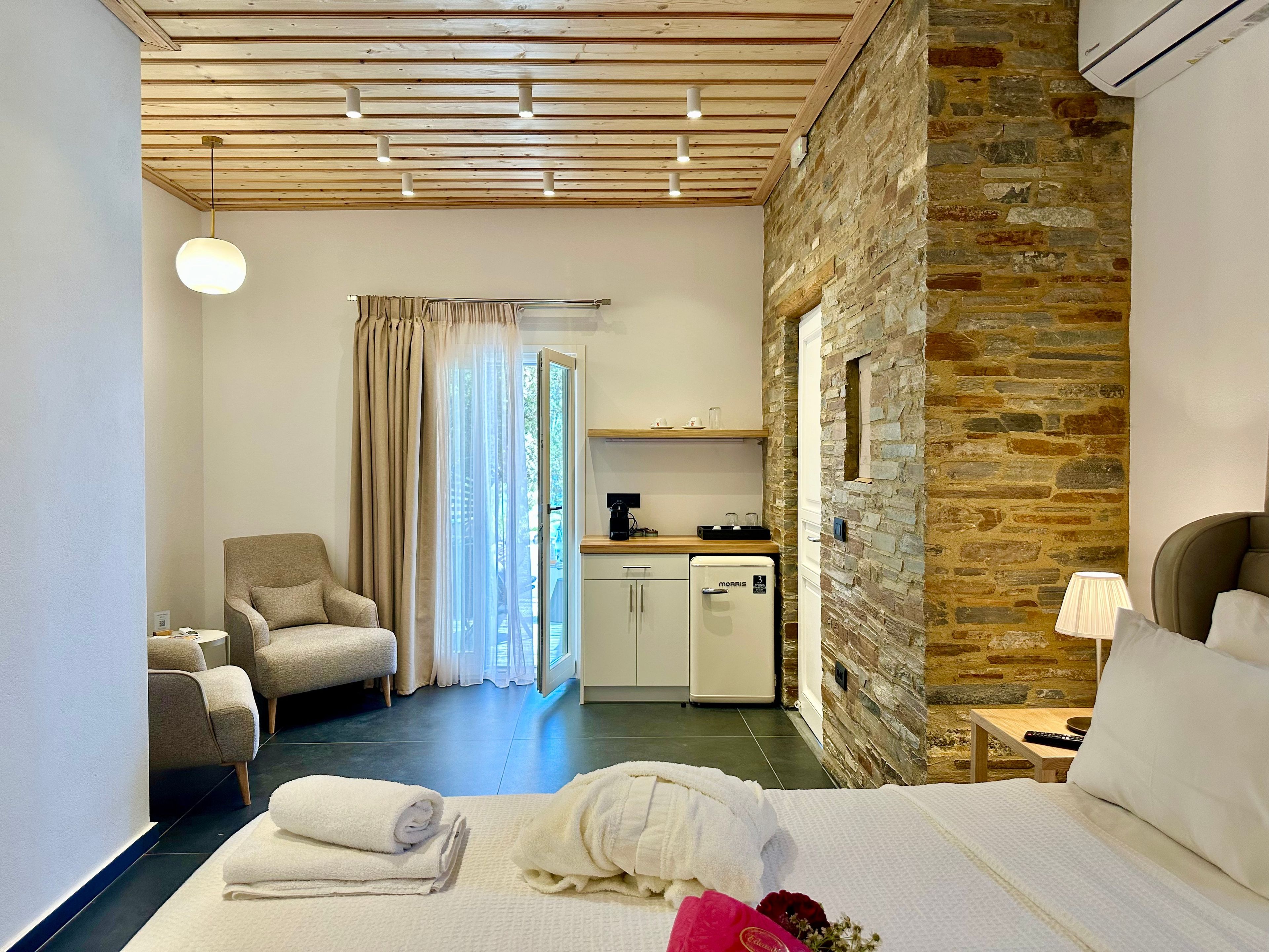 A hotel room with a bed, a chair, a desk with a coffee maker, a stone wall, and a window with curtains.