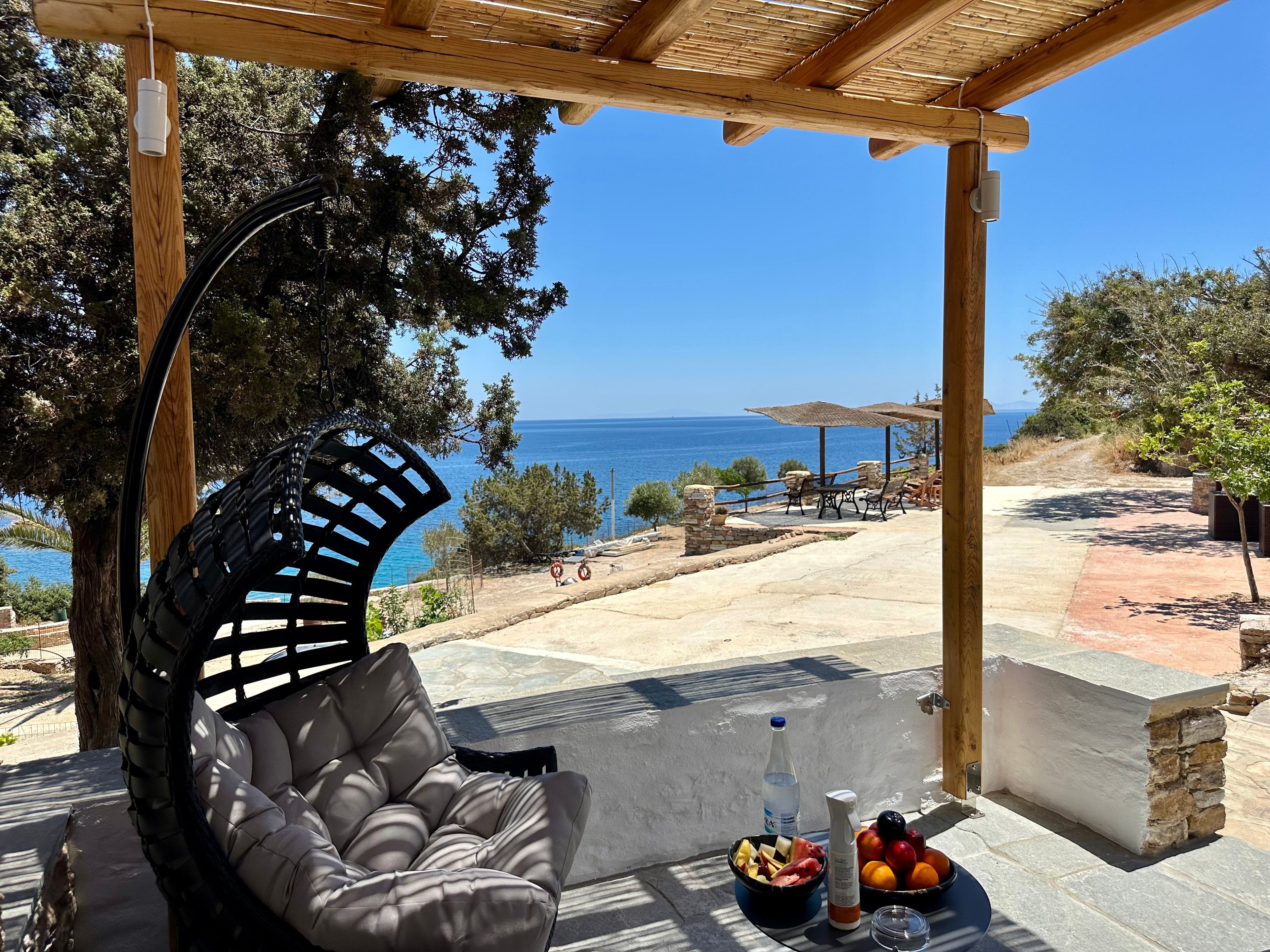 A wooden pergola with a cushioned chair and a table with fruit and a bottle. A clear view of the sea is visible through the trees.