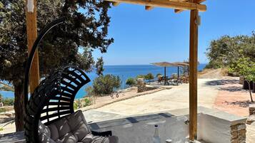 A wooden pergola with a cushioned chair and a table with fruit and a bottle. A clear view of the sea is visible through the trees.