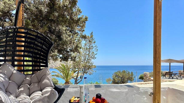 A wicker chair with a cushioned seat, a table with a fruit bowl and a bottle, and a view of the sea. - White & black Suites – Suite Five (Σίφνος)
