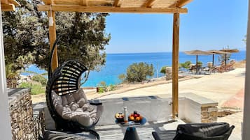 Ein überdachter Sitzbereich im Freien mit Blick auf das Meer, ein Hängekorbstuhl und ein Tisch mit Früchten und Getränken.