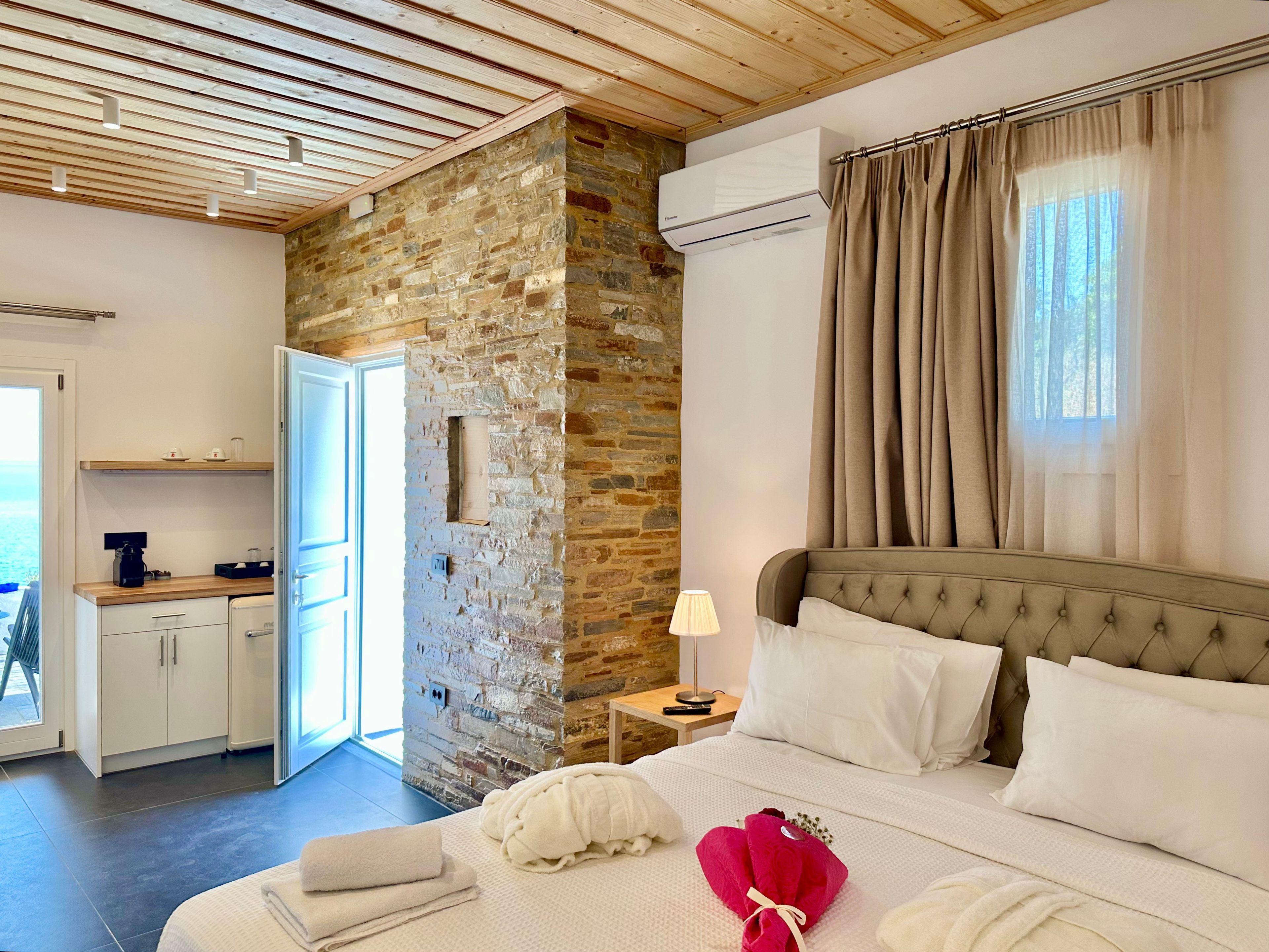 A hotel room with a stone wall, a bed with white bedding, a bedside table with a lamp, and a view of the sea through a window.