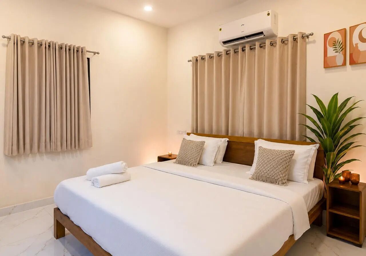 A hotel room with a bed, curtains, an air conditioning unit, a bedside plant, and artwork on the wall.
