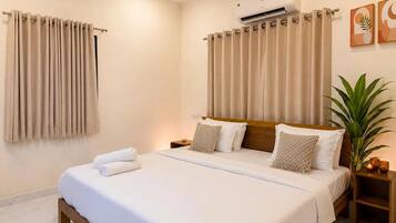 A hotel room with a bed, curtains, an air conditioning unit, a bedside plant, and artwork on the wall.