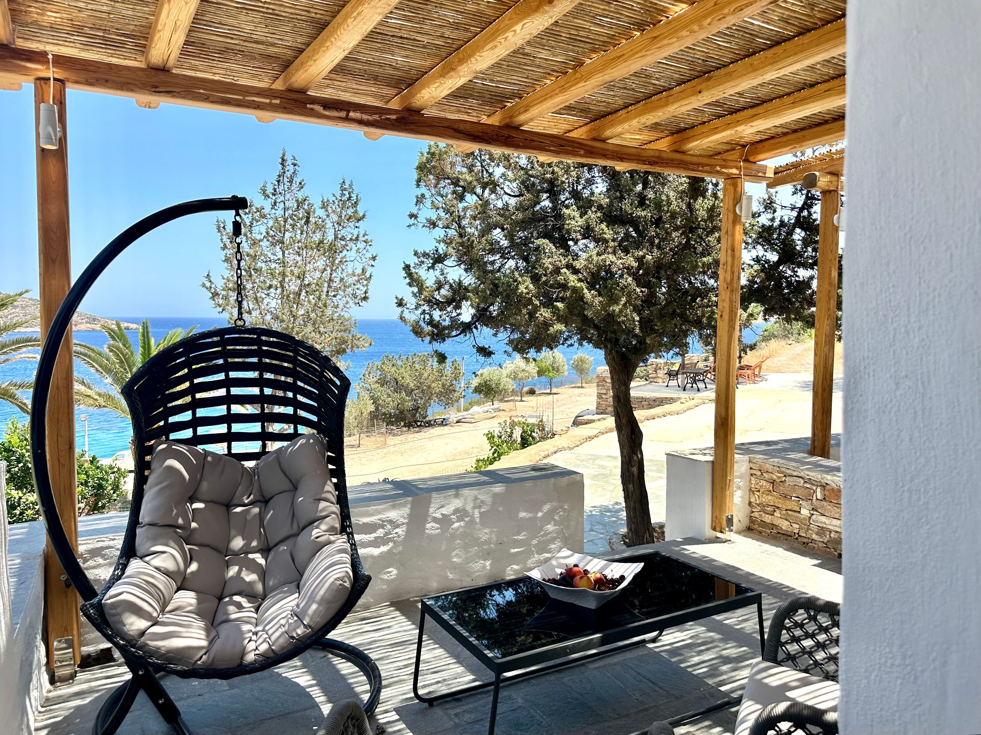 A covered outdoor seating area with a cushioned chair, a table with fruit, and a view of the sea.