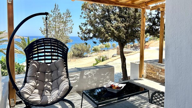 A covered outdoor seating area with a cushioned chair, a table with fruit, and a view of the sea. - White & black Suites – Suite Five (Platis Yialos Sifnos)