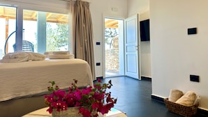 A bedroom with a bed, a window, a door, and a wall-mounted TV. - White & black Suites – Suite Five (Platis Yialos Sifnos)