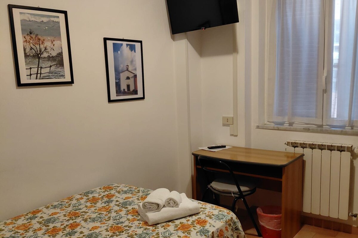 A bedroom with a bed, a desk, a chair, a television, and a window with curtains.