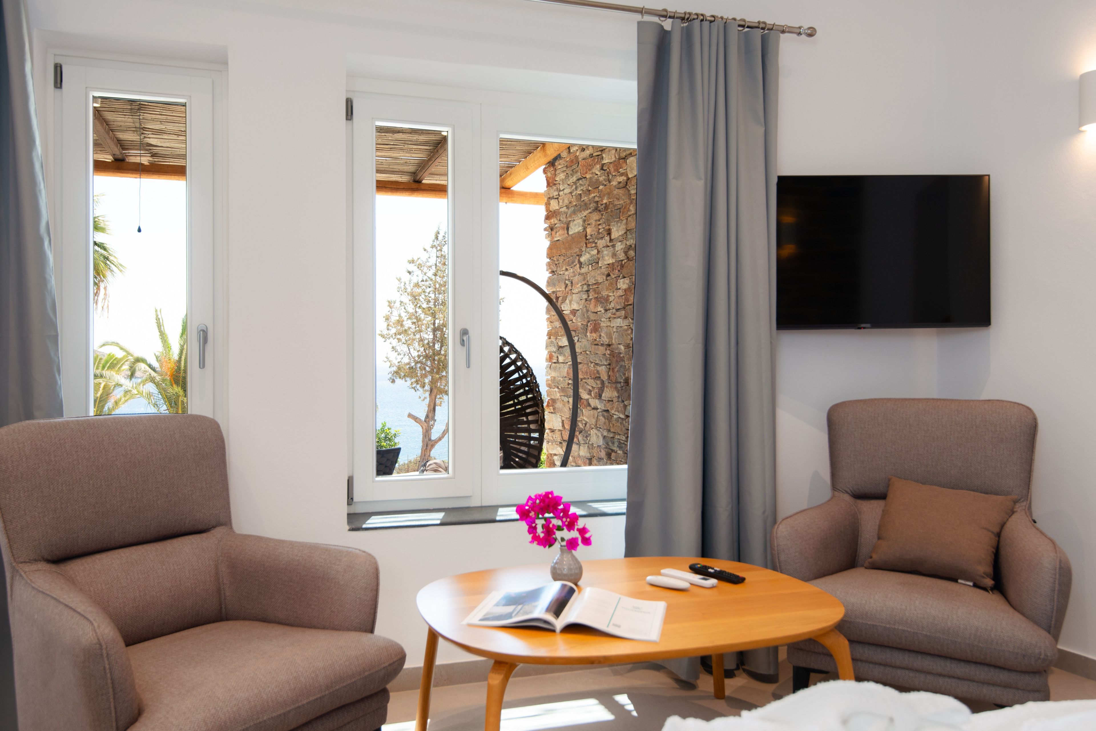Una habitación con dos sofás, una mesa con un libro y controles remotos, un televisor en la pared y un ventanal con vistas al mar y a los árboles.